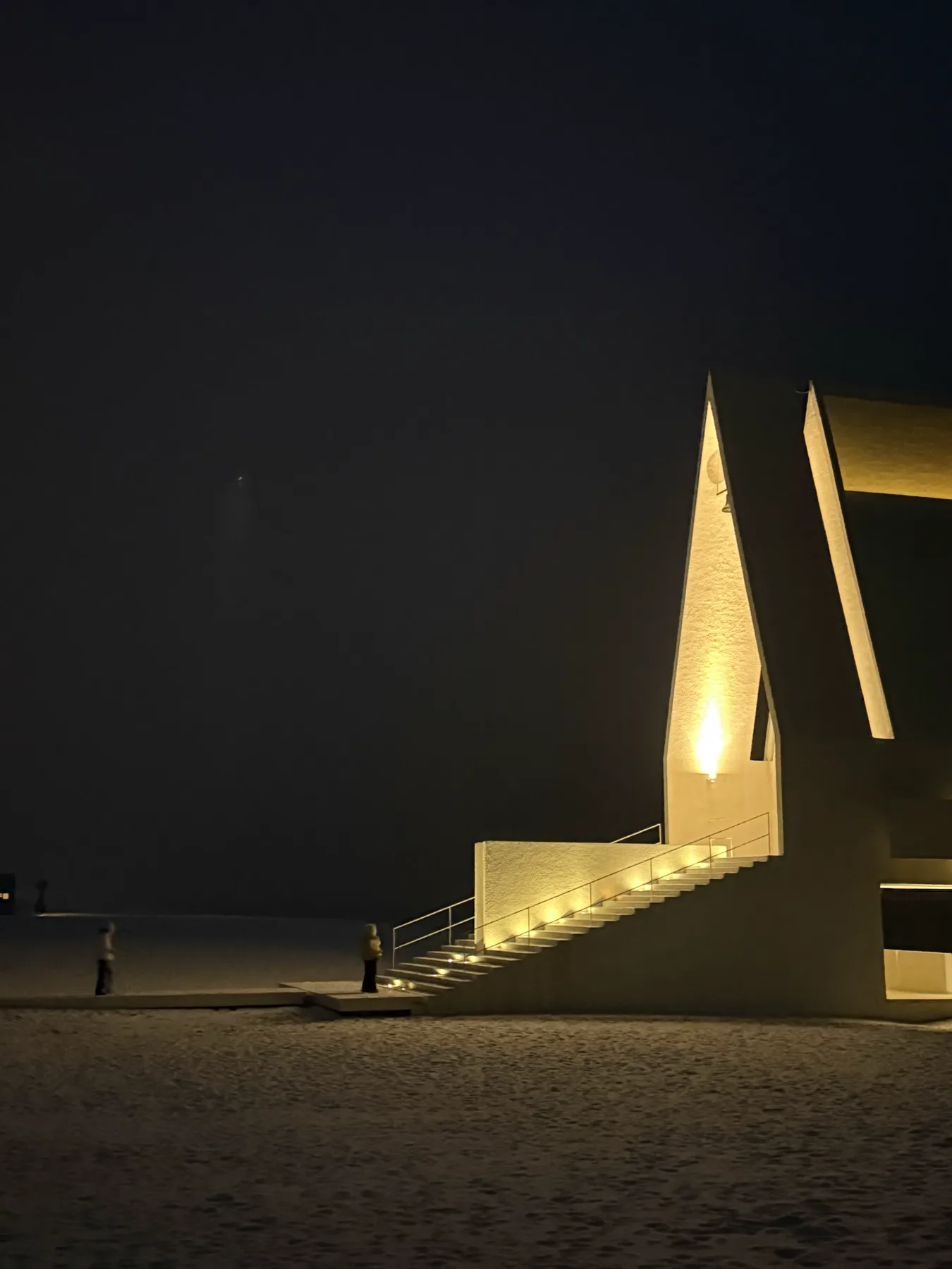 Original night photograph of two figures beside an illuminated angular house